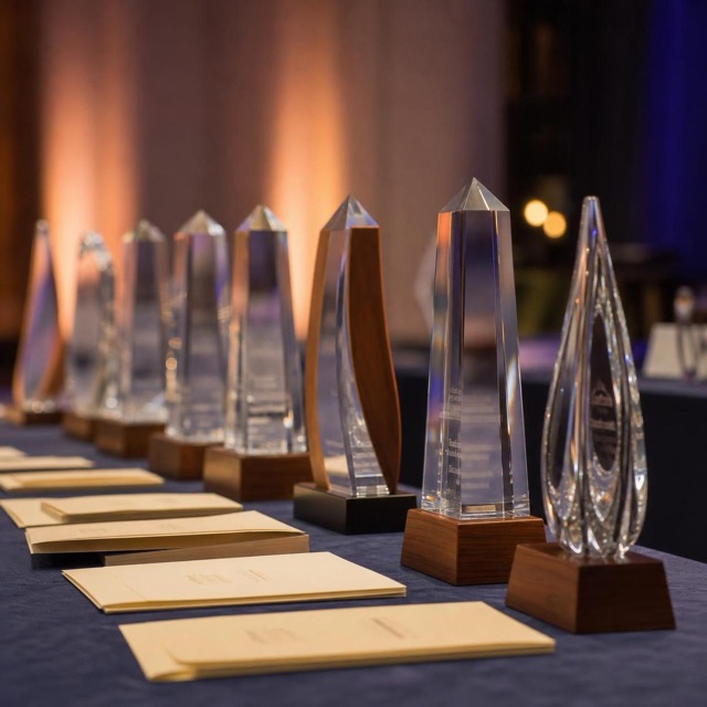 Row of crystal and koa-wood award trophies on a slate-blue table before a corporate gala