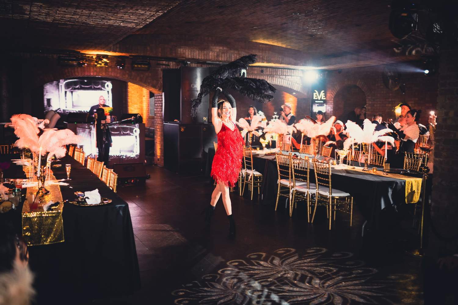 Long tables with feather centerpieces and a dancer in a red fringe dress at a Great Gatsby soirée produced by JJ Signature Experiences