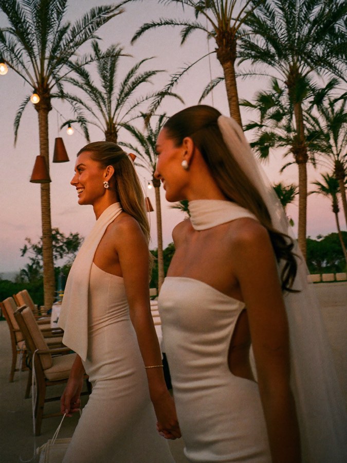 Two brides walking hand in hand beneath palms at dusk, seen in profile